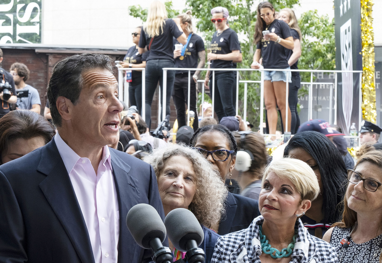 With members of the U.S. women's soccer team in the background before the start of a ticker tape parade in their honor, Gov. Andrew Cuomo speaks before signing a bill into law Wednesday, July 10, 2019, in New York. The bill will expand a law banning gender pay discrimination to make it illegal for employers to pay workers differently based on their age, race, religion or other characteristics, and making it easier for workers to prove pay discrimination in court. (AP Photo/Craig Ruttle)