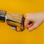 Bionic robot arm and the human arm are knocking fists, a greeting sign on a bright yellow background, interaction and rapprochement of a person with new modern technologies.