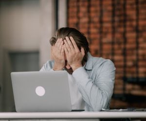 Business hand in front of laptop, with hands to face appearing to be stressed.