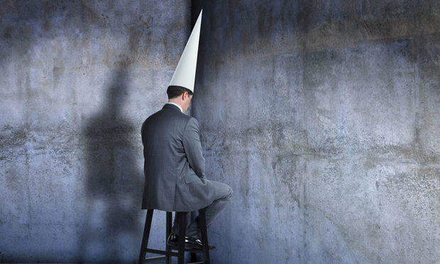 Bunsinessman in dunce cap sits on a stool in corner